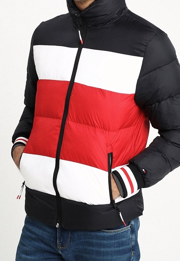 Uomo che indossa un piumino colorato con strisce orizzontali nere, bianche e rosse, con le mani nelle tasche con zip, abbinato a jeans blu.