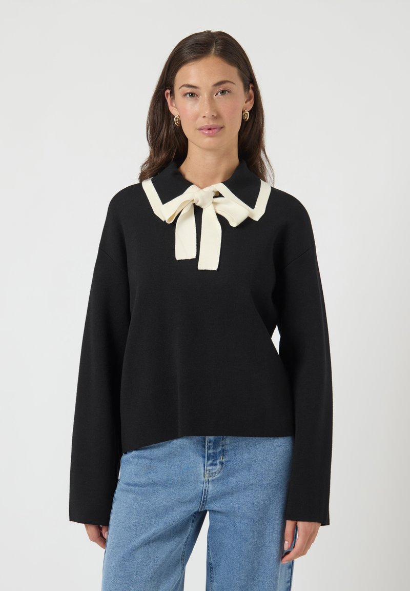 Kvinde iført en sort langærmet top med en stor hvid krave og sløjfe, kombineret med lyseblå jeans, står foran en ensfarvet baggrund.