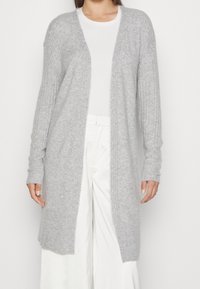 Cardigan grigio lungo con texture a coste e frontale aperto. Maniche lunghe e vestibilità rilassata. Indossato sopra un top bianco e abbinato a pantaloni bianchi.