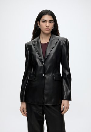 Frau mit langen dunklen Haaren, die einen schwarzen Lederblazer, ein bordeauxrotes Oberteil und schwarze Hosen trägt, steht vor einem einfarbig hellen Hintergrund.