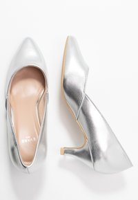 Chaussures à talons hauts argentées avec un design à bout pointu et ouvert sur les côtés. Texture lisse et brillante, talon en bois clair et intérieur rembourré.