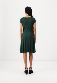 Robe verte mi-genou avec manches courtes, en tissu lisse. Présente une taille cintrée et une jupe évasée, associée à des sandales à talons épais noires.