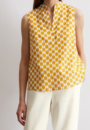 Blouse sans manches jaune moutarde à motifs géométriques blancs portée avec un pantalon blanc cassé, main gauche du mannequin dans la poche.