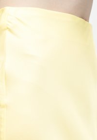 Krupni plan osobe koja nosi glatku, blijedo žutu suknju s visokim strukom na bijeloj pozadini.