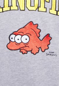 T-shirt gris arborant un poisson orange brodé à trois yeux, avec une texture subtile et une petite signature d'artiste intégrée dans le tissu.