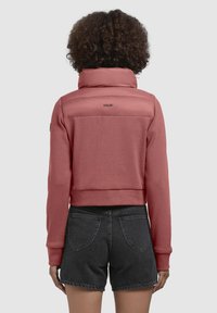 Roze jas met hoge kraag, textuur in de onderste stof en een glad bovenpaneel. Heeft een merkdetail op de achterkant. Gemaakt in combinatie met donkere shorts.