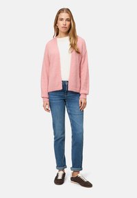 Roze pluizige cardigan met open voorkant, gedragen over een wit shirt. Lichtblauwe spijkerbroek en donkerbruine sneakers met witte veters maken de outfit compleet.