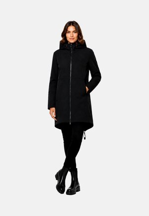 Woman wearing a black hooded coat with front zipper, black pants, and black ankle boots, standing with one hand in pocket and legs crossed.