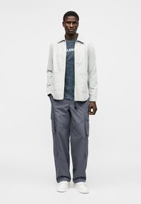 Άνδρας που φοράει ανοιχτό γκρι μπουφάν με φερμουάρ, σκούρο μπλε γραφικό μπλουζάκι, γκρι παντελόνια cargo και λευκά αθλητικά παπούτσια, στέκεται μπροστά σε λευκό φόντο.