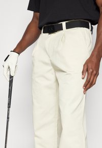 Λευκό βαμβακερό παντελόνι με πτυχές στο μπροστινό μέρος, συνδυασμένο με μια μαύρη πλεγμένη ζώνη με λογότυπο CK. Ένα λευκό γάντι γκολφ κρατά μια μαύρη γήινη ράβδο.