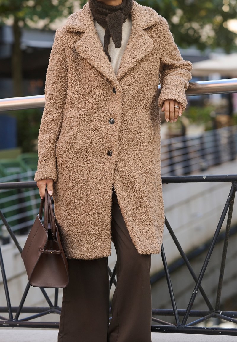 Personne portant un manteau beige texturé et une écharpe sombre, tenant un sac à main marron, debout près d'une rambarde en métal à l'extérieur.