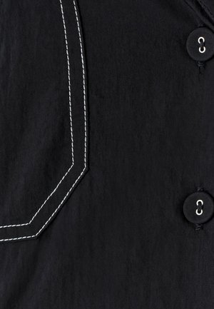 Czarna tkanina z białymi, przeszytymi kątowymi szwami oraz dwoma czarnymi guzikami z białą nitką zabezpieczonymi w dziurkach.