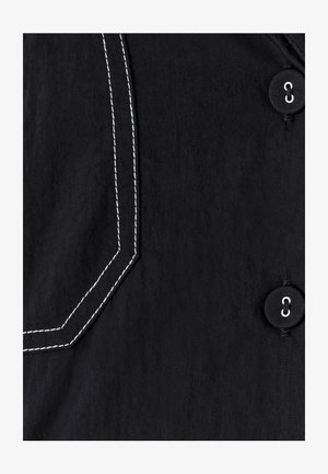 Czarna tkanina z białymi, przeszytymi kątowymi szwami oraz dwoma czarnymi guzikami z białą nitką zabezpieczonymi w dziurkach.