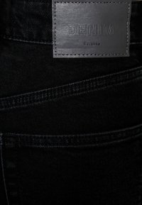 Tejido de denim negro con una textura característica. Cuenta con una etiqueta de cuero etiquetada como "DENIM" y "Bershka", con detalles de costura en contraste.