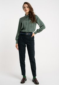 Zielona satynowa bluzka z kołnierzykiem, w połączeniu z ciemnymi spodniami w kratę, granatowym paskiem z złotym pierścieniem oraz brązowymi mokasynami i zielonymi skarpetkami.