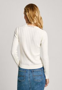 Biely žebrovaný sveter s dlhými rukávmi, ktorý má priliehavý strih a malé logo "Pepe Jeans" na zadnom výstrihu. V kombinácii s modrou riflovou sukňou.