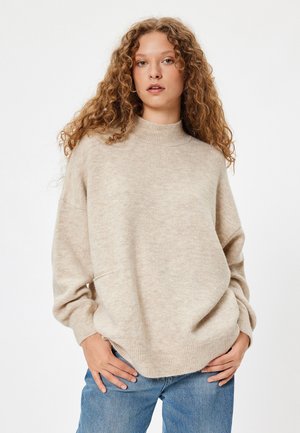 Beige gestrickter Pullover mit hohem Kragen, überschnittenen Schultern und gerippten Bündchen. Weiche Textur, leicht übergroßer Schnitt, kombiniert mit blauen Jeans.