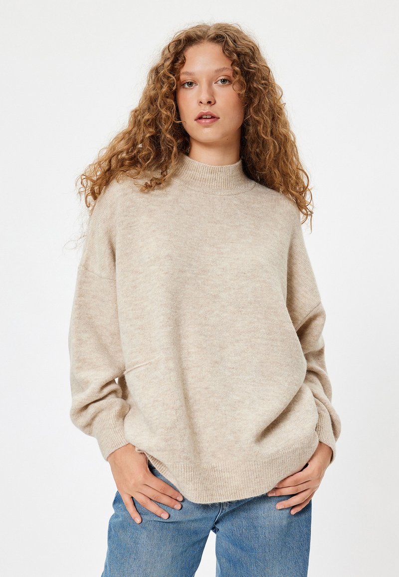 Beżowy sweter dziany z wysokim kołnierzem, opadłymi ramionami i prążkowanymi mankietami. Miękka tekstura, nieco oversize'owy krój, w zestawieniu z niebieskimi dżinsami.