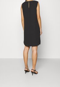 Robe noire sans manches avec un dos en trou de serrure, longueur genou, confectionnée en tissu lisse. Assortie à des sandales à talons noires.