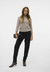 Pull en maille beige ajusté à manches trois quarts, associé à un pantalon noir bien taillé et des bottines brunes. Pochette noire tenue à la main.