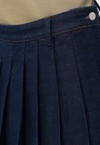 Tumes pitsidega denim seelik, millel on jäik vöökoht ja oranž lõng. Omab metallist nuppu ja vertikaalseid pitside tekstuuri lisamiseks.