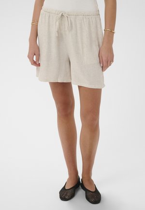 Žena oblečená v béžových ľanových šortkách so sťahovacou šnúrkou a čiernych sieťkových topánkach bez šnúrok, stojaca s rukami voľne pozdĺž tela.