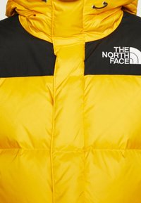 Veste doudoune jaune avec une section supérieure noire, design matelassé et logo sur la poitrine. Le matériau est lisse et brillant.