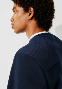 Marineblå genser med strukturelt mesh øverste del, glatt nedre stoff og hvit rund hals. Fokuserer på ergonomisk design.