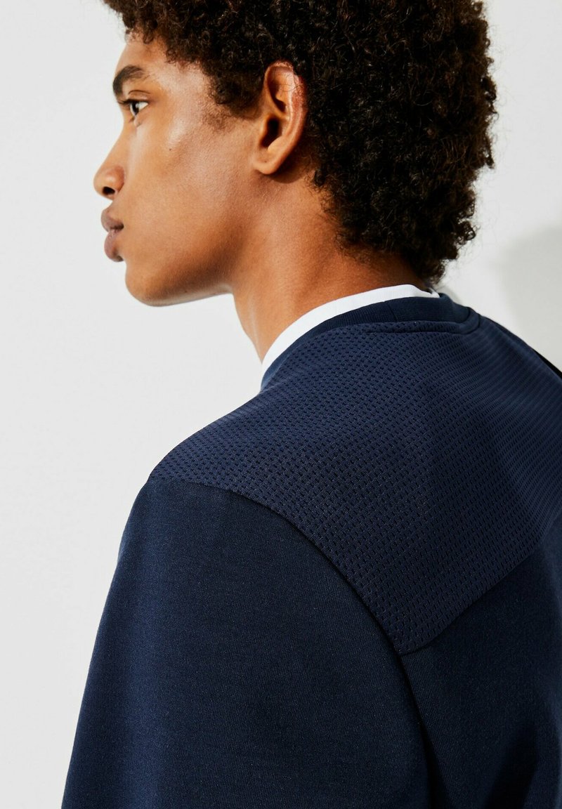 Sweatshirt bleu marine avec une partie supérieure en maille texturée, un tissu inférieur lisse et un col rond blanc. Accent sur le design ergonomique.