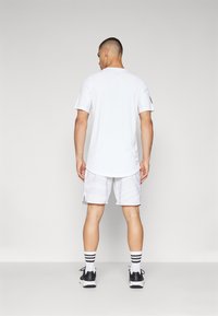 Weißes, kurzärmliges Sportshirt, kombiniert mit hellgrauen Shorts mit dezenten Mustern, getragen mit schwarzen Schuhen und gestreiften Socken.