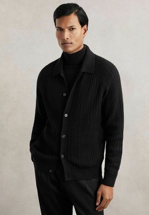 Mężczyzna w czarnym swetrze z dzianiny warkoczowej zapinanym na guziki, noszący czarny golf, stoi na tle jednolitego jasnoszarego tła.