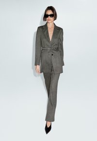Donna che indossa un completo grigio cintura con pantaloni, tacchi a punta neri e occhiali da sole neri su uno sfondo chiaro uniforme.