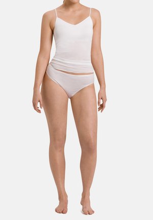 Vrouw staat blootsvoets, draagt een wit hemdje en bijpassende witte bikini-onderbroek tegen een effen achtergrond.