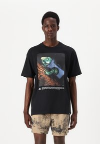 Sort t-shirt med et farverigt iguana-grafik på fronten, med fed tekst nedenfor. Stoffet ser blødt og lidt tekstureret ud.