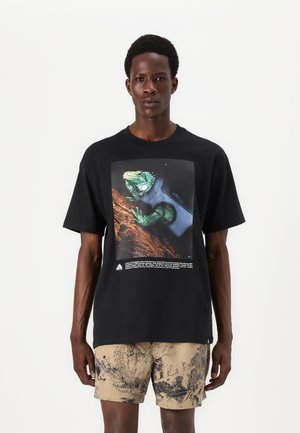 Schwarzes T-Shirt mit einem bunten Leguan-Graphic auf der Vorderseite, darunter in fetter Schrift. Der Stoff wirkt weich und leicht strukturiert.