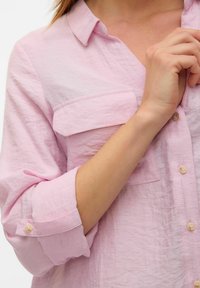 Camisa de botones de color rosa claro con cuello, que presenta un bolsillo frontal, textura suave, mangas enrolladas y pequeños botones de plástico.