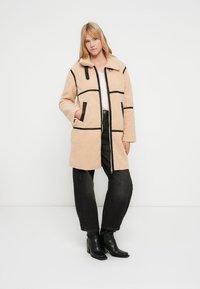 Beige fleece jas met zwarte biezen, voorzien van een kraag, voorzakken en een ritssluiting. Gecombineerd met zwarte wijde spijkerbroek en enkellaarsjes.