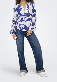 Blusa bianca con motivi floreali blu, scollatura a V e maniche lunghe a sbuffo, abbinata a jeans a gamba larga blu scuro e sneakers bianche.