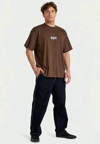 Homem de pé com uma mão no bolso, vestindo uma t-shirt castanha da Billabong, calças escuras com costura branca e sapatos pretos.