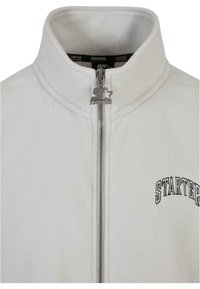 Valkoinen fleece-takki, jossa on korkea kaulus, hopea vetoketju ja musta kirjailtu "STARTER"-logo vetoketjun yläpuolella.