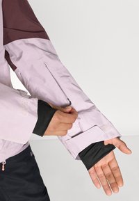 Vaalean lila ulko-osa takin hiha, jossa on viininpunainen korostus. Takissa on vetoketjullinen tasku ja musta sisätila. Se on valmistettu sileästä, vettä hylkivästä materiaalista.