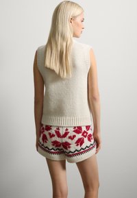 Strickweste in Creme mit weiten Ärmeln, kombiniert mit cremefarbenen Shorts mit roten Blumenmustern und schwarzen Akzenten. Die Textur ist dick und kuschelig.