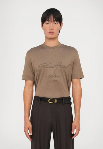 Camiseta corta de manga corta en color marrón con un ajuste relajado, que presenta detalles bordados en forma de texto. Se lleva con pantalones oscuros y un cinturón negro.