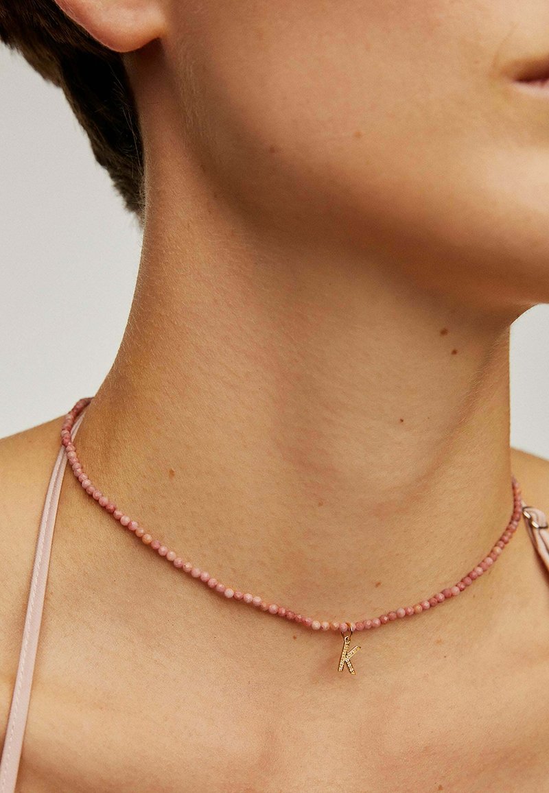 Collar choker de cuentas rosas con pequeñas cuentas redondas y un colgante en forma de letra "K" dorada, reposando sobre el cuello contra una piel suave.