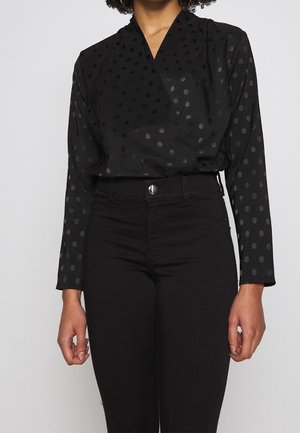 Zwarte blouse met een V-hals en lange mouwen, met een ton-sur-ton polkadotpatroon, gecombineerd met zwarte jeans met hoge taille.