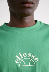 Camiseta de algodón verde con un clásico cuello redondo y un logotipo bordado de "ellesse" en crema, centrado en el pecho.