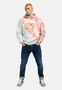 White hoodie with a pink and yellow swirled pattern on one side, featuring a red "Coca-Cola" logo, paired with dark jeans and patterned sneakers.