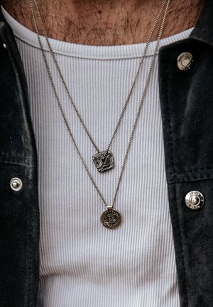 Nahaufnahme einer Person, die ein weißes, geripptes Hemd und eine schwarze Jacke mit zwei silbernen Halsketten trägt, eine mit quadratischem und eine mit rundem Anhänger.