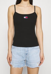 Czarny ribbed top na ramiączkach spaghetti, z małym haftowanym logo w czerwonym, białym i niebieskim kolorze na piersi. W komplecie z jasnoniebieskimi szortami dżinsowymi.