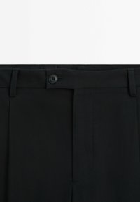 Section de la taille d'un pantalon noir habillé avec fermeture à bouton, passant de ceinture et braguette zippée cachée sur fond blanc.